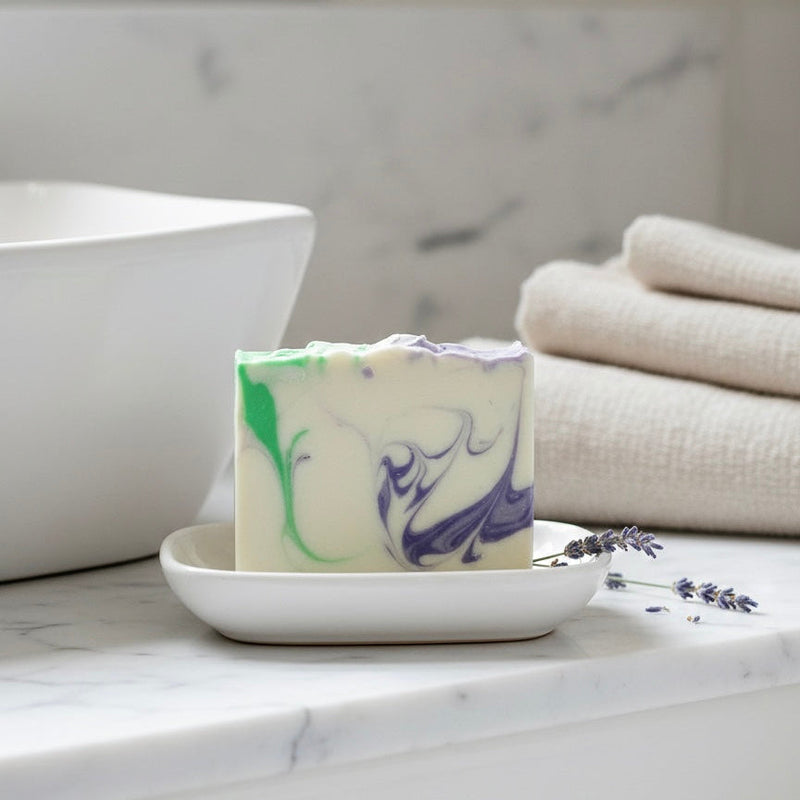 Finishya lavender and green marbled bar soap sitting on a white ceramic dish in a marble bathroom, next to sprigs of fresh lavender and white towels.