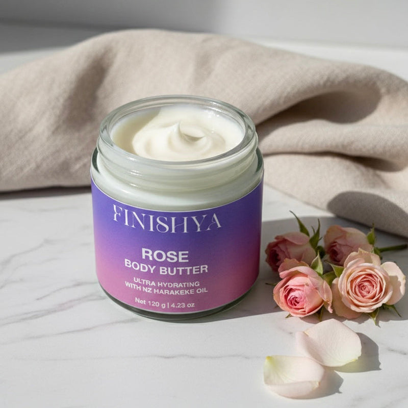 An open jar of Finishya Rose Body Butter sits on a marble surface, surrounded by pink rose buds and petals. A lifestyle image showcasing the vegan, Harakeke-infused body butter made in NZ.