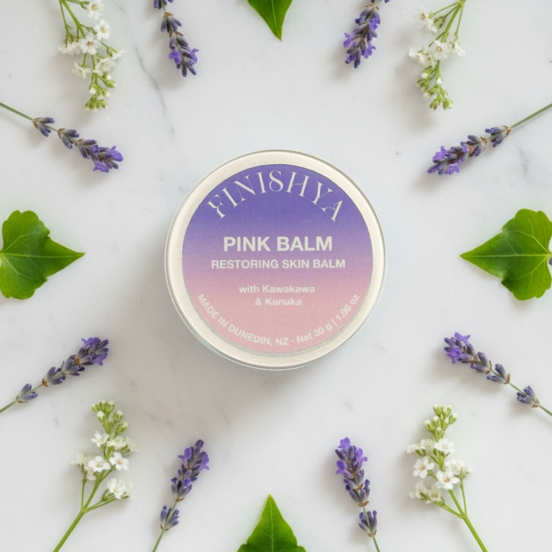 Finishya Pink Balm tin centered in a botanical flat lay with lavender and green leaves, showcasing the cruelty-free ingredients used for skin restoration.