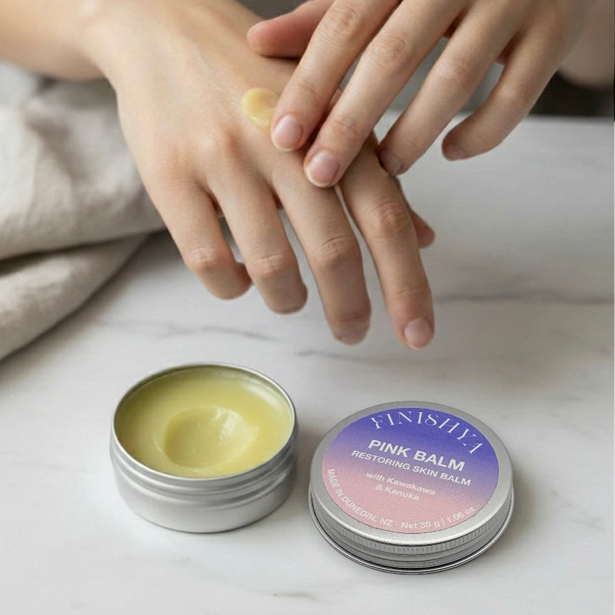 Person applying Finishya pink kanuka balm to hand with balm container and lid on a light surface.