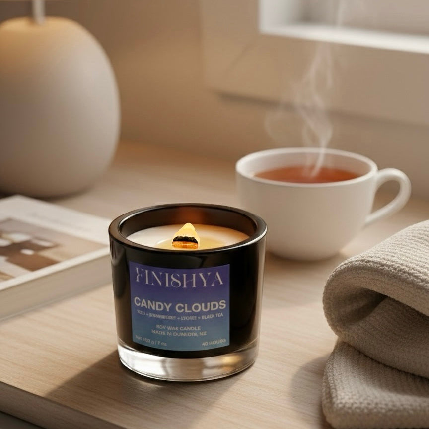 A lit Finishya Candy Clouds wood wick candle on a wooden table next to a cup of tea, creating a cozy morning atmosphere with soft steam.