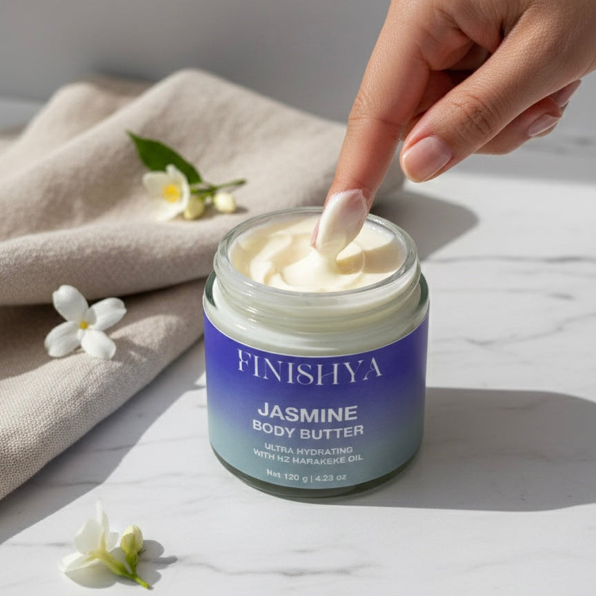 A close-up of a finger dipping into a jar of Finishya Jasmine Body Butter, showing the rich, white cream. The jar features a blue-to-teal gradient label on a marble surface with jasmine flowers.
