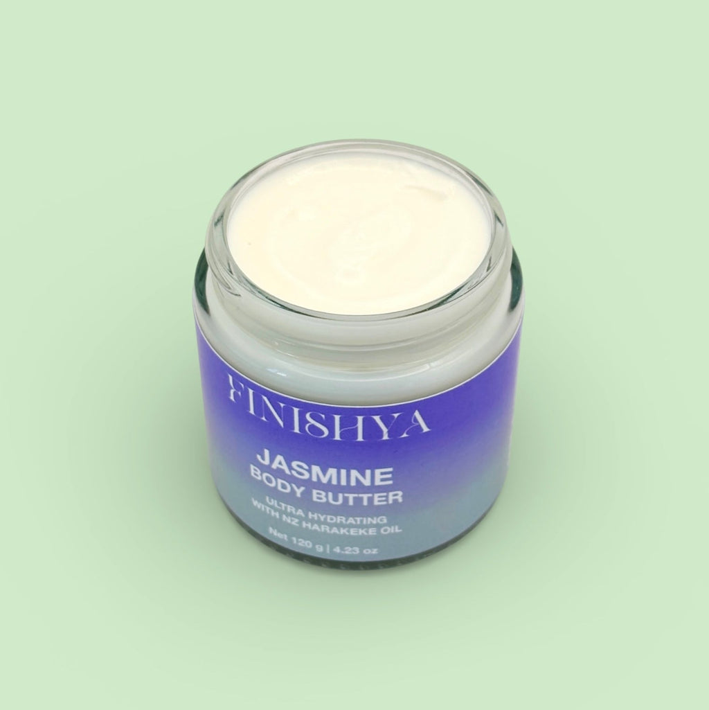 High-angle view of an open jar of Finishya Jasmine Body Butter, showcasing the thick, creamy consistency of the moisturiser. The jar sits against a minimalist light green background.