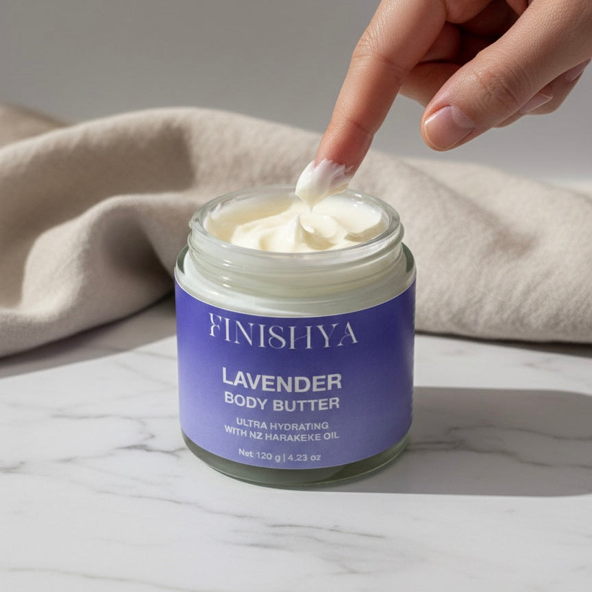 A close-up of a finger dipping into a jar of Finishya Lavender Body Butter, showing the thick, creamy white texture. The jar features a purple label and sits on a marble surface with a beige linen cloth.