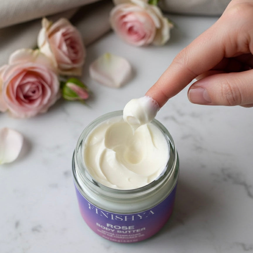 A close-up of a finger scooping out Finishya Rose Body Butter from a jar. The creamy texture is visible against a marble background with fresh pink roses and a beige linen cloth.