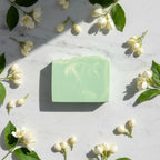 Overhead shot of a green marbled Jasmine soap bar surrounded by white jasmine blooms and green leaves on a marble surface.