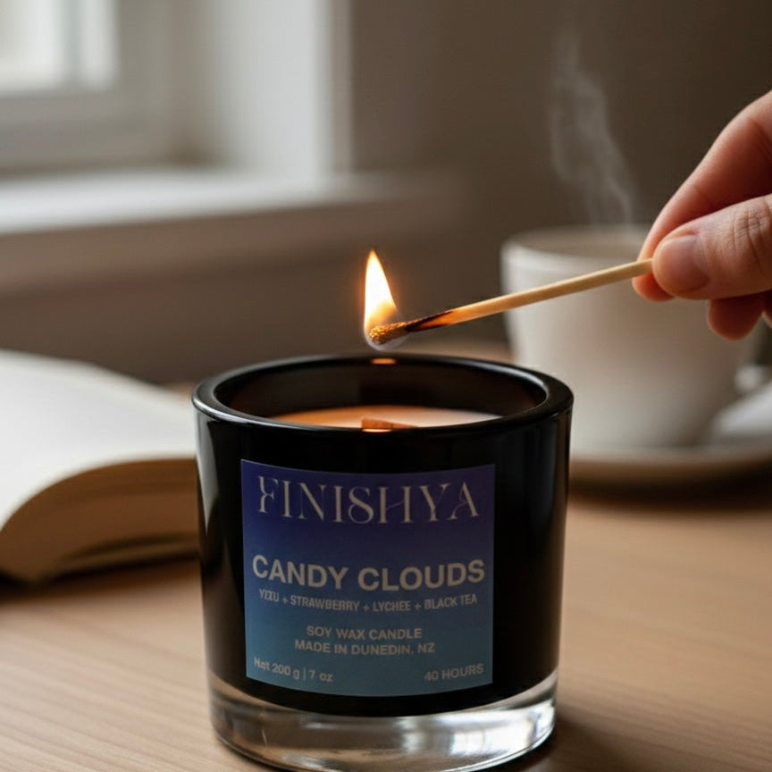 Close-up of a hand lighting a Finishya Candy Clouds soy wax candle with a match, highlighting the crackling wood wick and luxury black glass vessel.