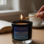 Close-up of a hand lighting a Finishya Candy Clouds soy wax candle with a match, highlighting the crackling wood wick and luxury black glass vessel.