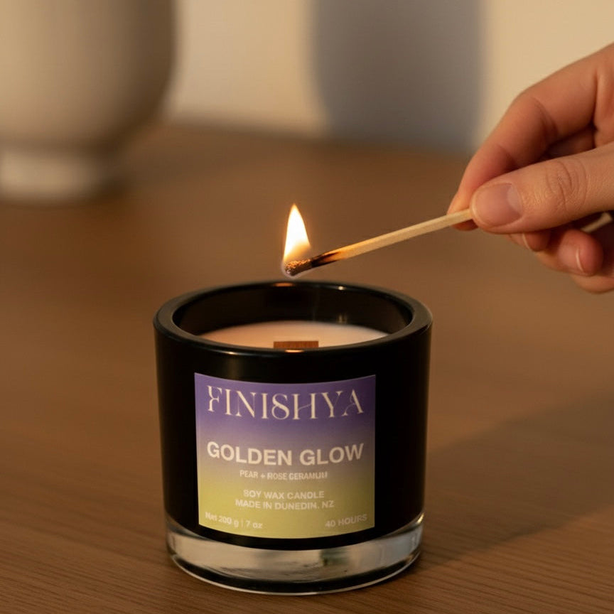 A hand lighting the wooden wick of a Finishya Golden Glow soy candle with a match in a cozy, warm-toned room.