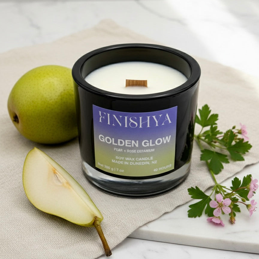 A black glass soy candle with a wooden wick sitting on a marble surface next to a fresh sliced pear and pink geranium flowers.