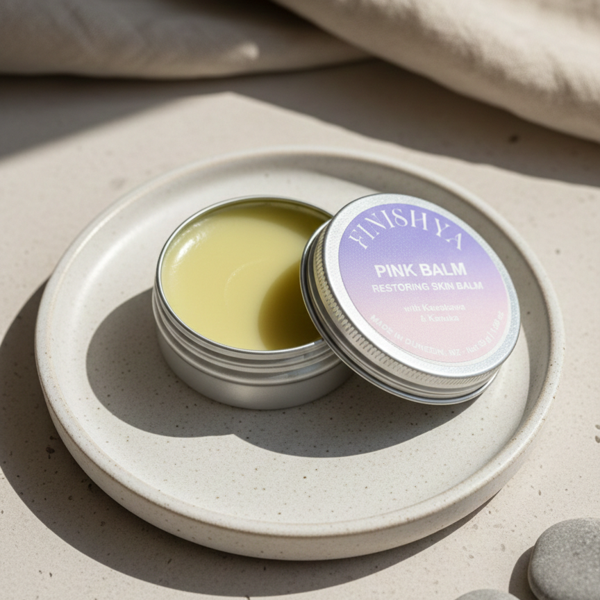 An open tin of Finishya Pink Balm resting on a ceramic plate in soft sunlight, displaying the kawakawa-infused balm texture.