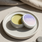 An open tin of Finishya Pink Balm resting on a ceramic plate in soft sunlight, displaying the kawakawa-infused balm texture.